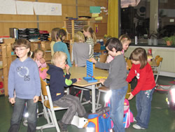 Eine Gruppe von Kindern in einem Raum, der wie ein Klassenzimmer oder Spielzimmer aussieht. Sie stehen um einen Tisch herum, und einige spielen ein Spiel, das wie „Vier gewinnt“ aussieht. Andere stehen oder sitzen in der Nähe und schauen zu oder beschäftigen sich mit anderen Dingen. Im Hintergrund sind verschiedene Schulsachen und Spielzeug zu sehen.