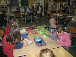 In einem Klassenzimmer sitzen vier Kinder an einem Tisch. Sie arbeiten oder lernen. Auf dem Tisch liegen Hefte und Stifte. Im Hintergrund sind weitere Kinder an Tischen zu sehen. An der Wand hängen Tafeln.