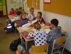 Kinder sitzen an Tischen in einem Klassenzimmer und arbeiten an Aufgaben. Auf den Tischen liegen Papiere, Stifte und Bücher. Ein Kind hält ein großes Blatt Papier in den Händen, während andere schreiben oder lesen.