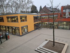 Ein Schulhof, der von einem modernen Gebäude mit gelben und roten Fassaden umgeben ist. In der Mitte des gepflasterten Hofes steht ein Baum in einer quadratischen, erhöhten Rabatte aus Beton. Es gibt Stufen, die zu einem Eingang führen, und große Fensterfronten. Im Hintergrund sind weitere Bäume und ein Teil eines anderen Gebäudes zu sehen. Der Himmel ist blau und wolkenlos. GGS Südschule Krefeld