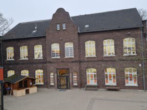 Backsteingebäude im Stil der Backsteingotik mit zwei Stockwerken und Sprossenfenstern. Der zentrale Giebel und der gepflasterte Hof mit einer kleinen Holzhütte prägen das Bild.