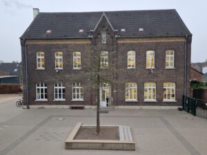 Historisches, zweistöckiges Backsteingebäude, eine alte Schule in Krefeld, mit Efeubewuchs an der linken Fassadenseite. Im Vordergrund befindet sich ein gepflasterter Schulhof mit einem jungen Baum und einer Bank.