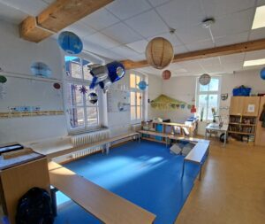 Heller Klassenraum mit blauem Teppich, Papierlaternen, Leseecke und Regalen