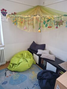 Kuschelige Leseecke mit Sitzkissen, Bücherregal und bunter Lichterkette unter Stoffhimmel