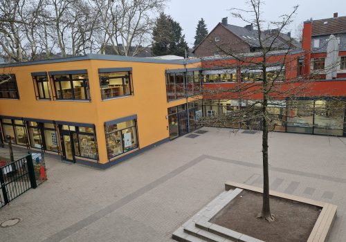 Ein Schulhof, der von einem modernen Gebäude mit gelben und roten Fassaden umgeben ist. In der Mitte des gepflasterten Hofes steht ein Baum in einer quadratischen, erhöhten Rabatte aus Beton. Es gibt Stufen, die zu einem Eingang führen, und große Fensterfronten. Im Hintergrund sind weitere Bäume und ein Teil eines anderen Gebäudes zu sehen. Der Himmel ist blau und wolkenlos. GGS Südschule Krefeld