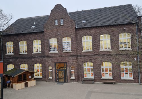 Backsteingebäude im Stil der Backsteingotik mit zwei Stockwerken und Sprossenfenstern. Der zentrale Giebel und der gepflasterte Hof mit einer kleinen Holzhütte prägen das Bild.