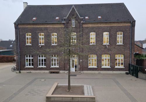 Historisches, zweistöckiges Backsteingebäude, eine alte Schule in Krefeld, mit Efeubewuchs an der linken Fassadenseite. Im Vordergrund befindet sich ein gepflasterter Schulhof mit einem jungen Baum und einer Bank.