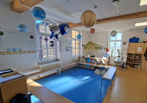 Heller Klassenraum mit blauem Teppich, Papierlaternen, Leseecke und Regalen