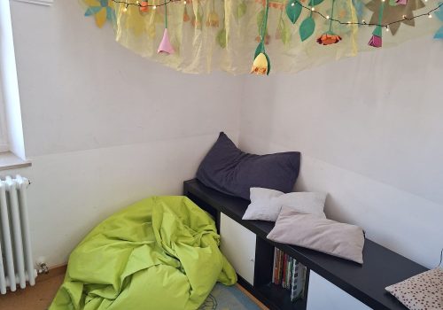 Kuschelige Leseecke mit Sitzkissen, Bücherregal und bunter Lichterkette unter Stoffhimmel
